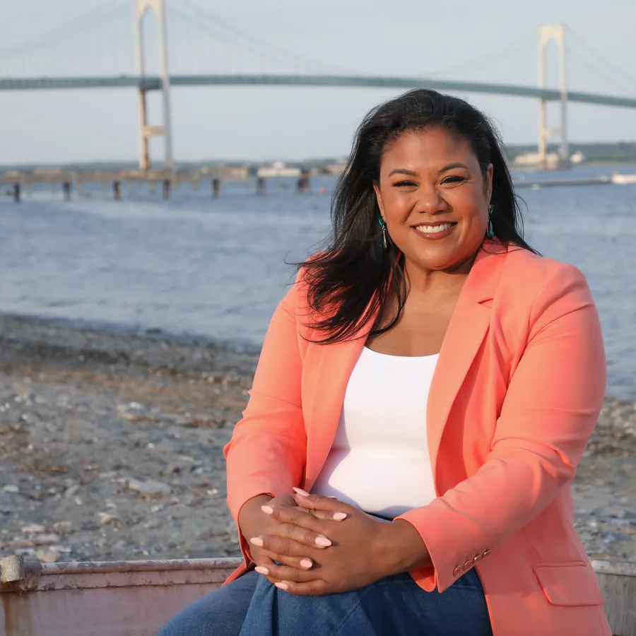 Angela Lima seated on the shoreline with the Claiborne Pell Newport Bridge behind her