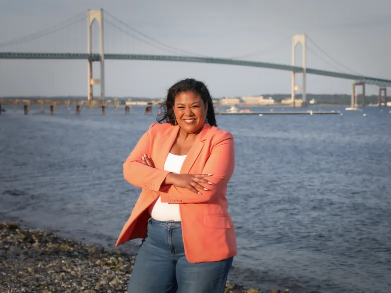 Angela Lima standing on the rocky shoreline with the Newport Bridge and Narragansett Bay behind her